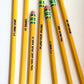 Six yellow pencils arranged diagonally on a plain white background, each engraved with a motivational phrase and featuring pink erasers with green and yellow horizontal striped bands.
