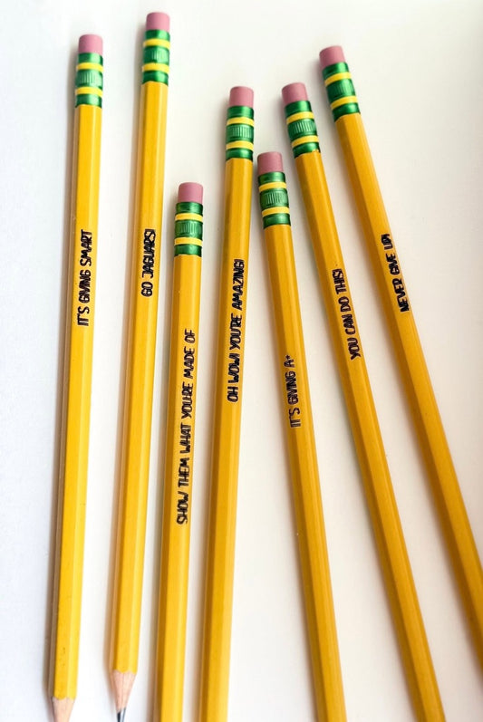 Six yellow pencils arranged diagonally on a plain white background, each engraved with a motivational phrase and featuring pink erasers with green and yellow horizontal striped bands.